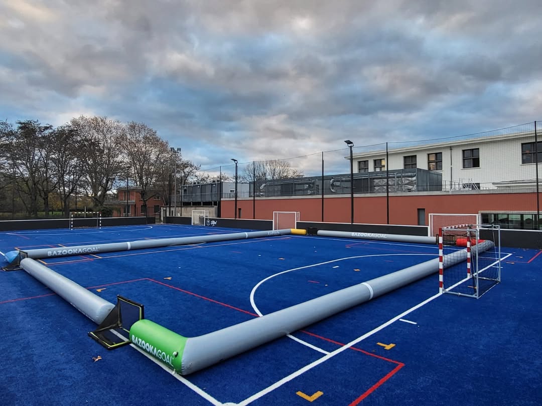 Campo de fútbol 3v3 AirPitch de la marca BazookaGoal con césped artificial azul y perímetros hinchables grises. Se ve una portería pequeña en primer plano y otras al fondo en una instalación deportiva al aire libre bajo un cielo nublado.